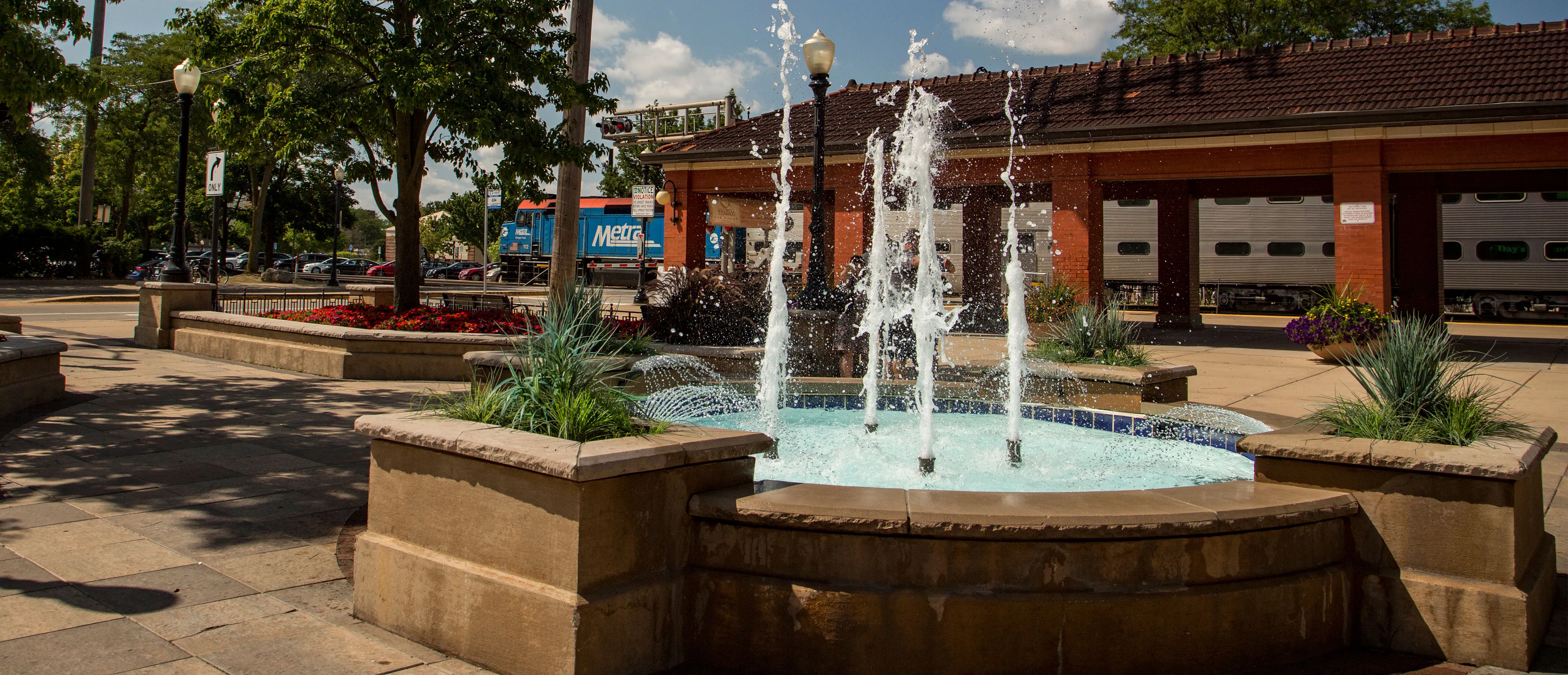 The famous fountain located in downtown Downers Grove pumps, water several feet into the air out of five visible jets. Shadows of shrubbery appear on the left, with trees visible in the back next to a vacant parking lot with brick awning clouds appear in the sky blue sky and a train traverses the horizon in the distance.