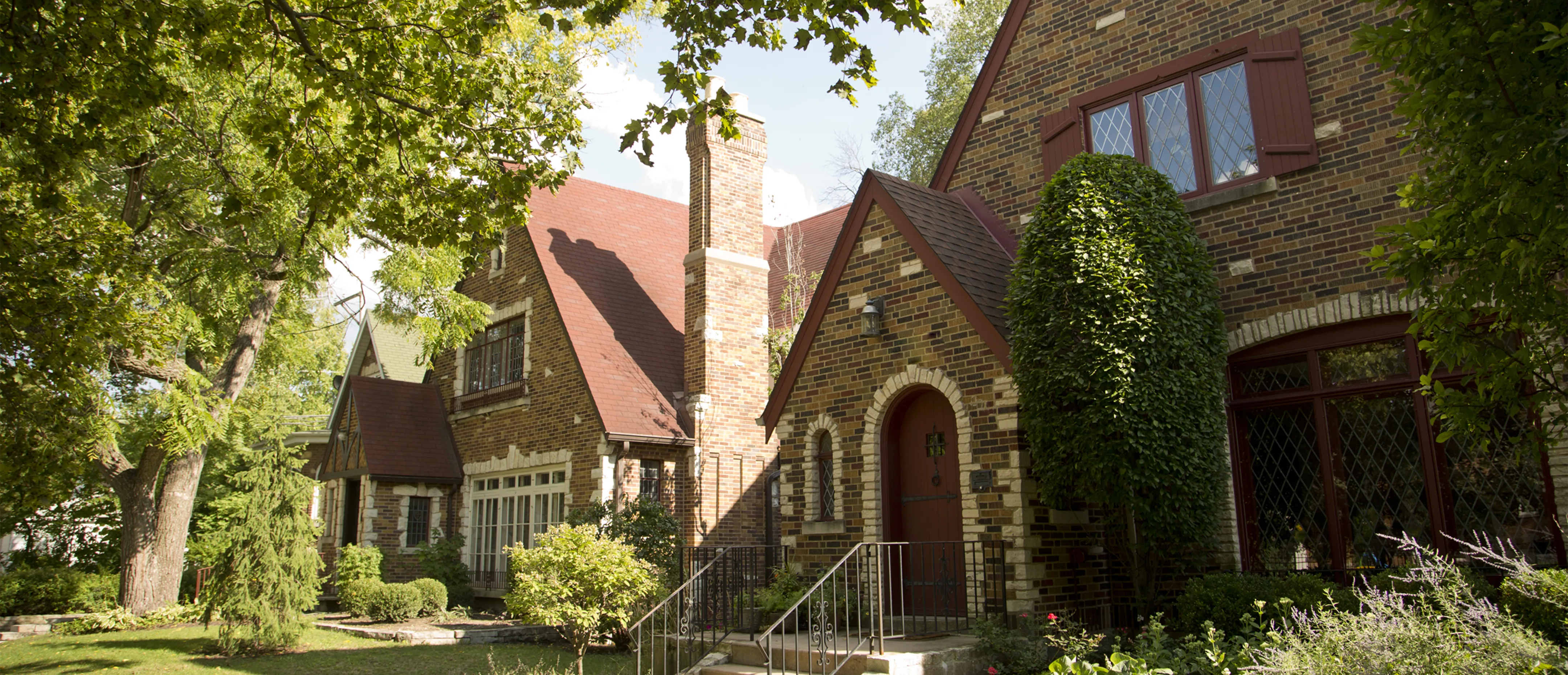 Gothic home features textured dark, brick, framing, dark, red, paneling and cream, colored brick accents lining cylindrical front door, first floor window, and single car garage. adjacent left lot contains shadowed three-story Family home with tall chimney. Sky is bright and various shades of green contrast against the dark façade of residential properties.