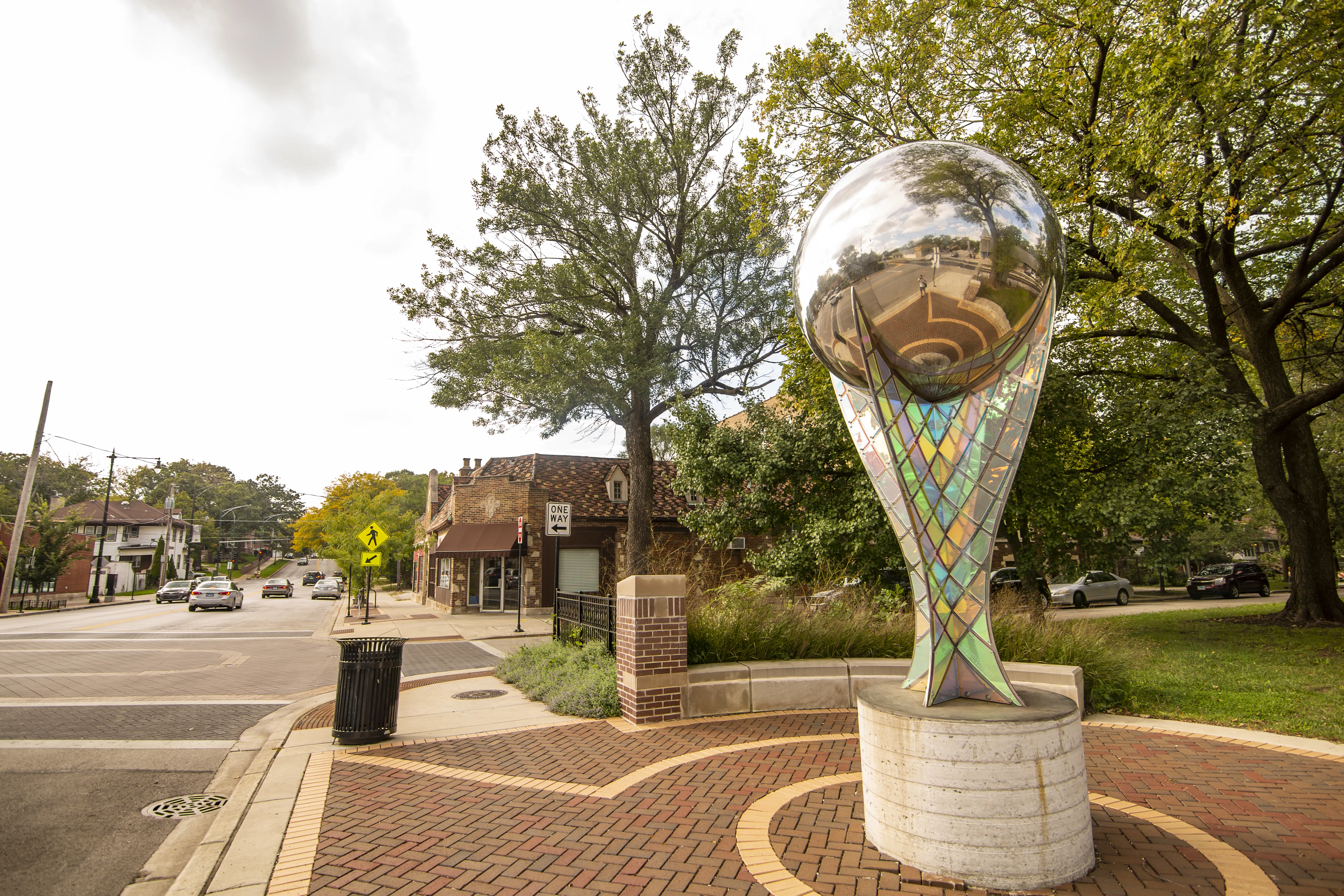 Statue with large spherical, mirror marble and colorful mural base sits in the right hand corner on the front lawn of downtown street. commercial and residential buildings appear on the horizon, and cars move towards the frame under a white sky lined with telephone wires and green leaves.